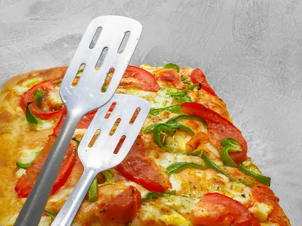 Pizza with tomato and green pepper. Fresh baked piece of pizza and tongs on the top, on grey background.