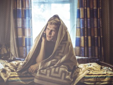 Genç adam yatağında telefon ile battaniye altında oturan yas