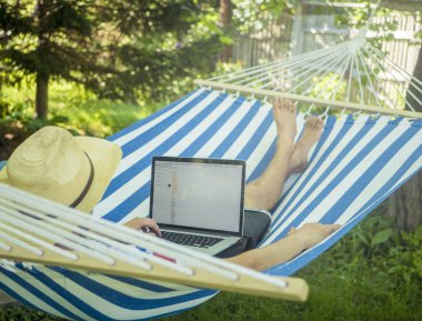 Bahçedeki açılan dizüstü bilgisayar ile hamakta yaz şapkalı rahatlatıcı delikanlı