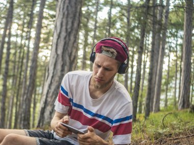 hipster içinde rahat ve kap oturup telefon ve smartphone üzerinde oyun oynamak kulaklık ile doğa ormandaki çılgın sakallı
