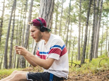 hipster içinde rahat ve kap oturup telefon ve smartphone üzerinde oyun oynamak kulaklık ile doğa ormandaki çılgın sakallı
