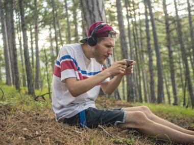 hipster içinde rahat ve kap oturup telefon ve smartphone üzerinde oyun oynamak kulaklık ile doğa ormandaki çılgın sakallı
