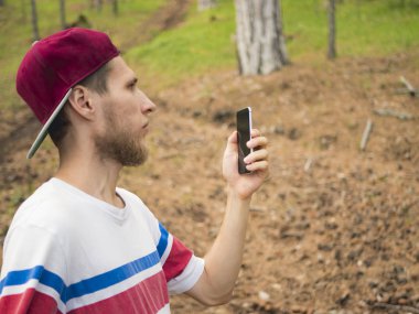 Genç sakallı adam İsviçre çam ormanda yürümek telefon ile fotoğraf çekme kapatın