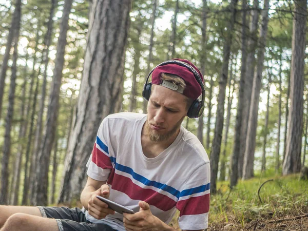 hipster içinde rahat ve kap oturup telefon ve smartphone üzerinde oyun oynamak kulaklık ile doğa ormandaki çılgın sakallı