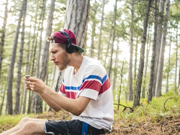 hipster içinde rahat ve kap oturup telefon ve smartphone üzerinde oyun oynamak kulaklık ile doğa ormandaki çılgın sakallı