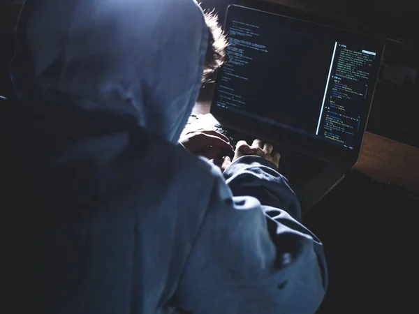 hacker in hoodie typing code in program using laptop in dark studio