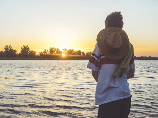 Вид ззаду молода людина з капелюхом і рушником, дивлячись на захід сонця, стоячи на пляжі — стокове фото