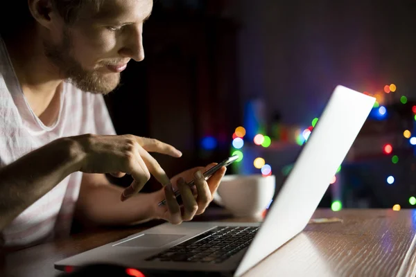 Joven hombre chat en red social con amigos utilizando el teléfono y el ordenador portátil tarde en la noche d — Foto de Stock