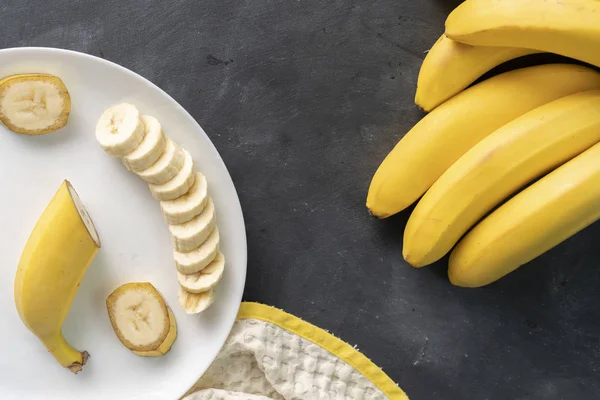 Banana fresca fatiada em uma placa de cerâmica branca na mesa preta, preparando ingredientes para pequenos-almoços saudáveis — Fotografia de Stock