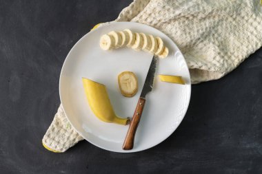 siyah masa üzerinde beyaz bir seramik tabakta taze dilimlenmiş muz, sağlıklı kahvaltı için malzemeler hazırlanıyor