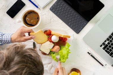 Genç erkek çalışırken laptopun önünde yemek yiyor, kötü alışkanlık ve obezite kavramları