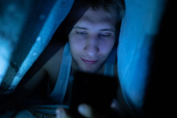 Nuori poika käyttää älypuhelinta makaa sängyssä peiton alla yöllä — kuvapankkivalokuva