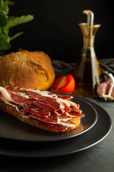 黒プレートと素朴な背景にイベリアハムとトーストパン 背景職人のパンで 新鮮なトマトとオリーブオイル — ストック写真