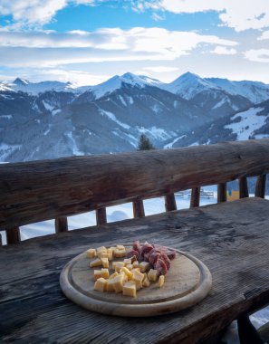 Wooden board with cheese and sausage on a balcony in front of snow covered mountains in the Austrian alps in winter clipart