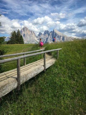 Alpe di Siusi - Seiser Alm ve Sassolungo - Langkofel dağ grubu mavi gökyüzünün önünde bulutlu. Dolomitler, Trentino Alto Adige, Güney Tyrol, İtalya 'da yaz aylarında iki çocuklu tahta ayaktakımı kayak merkezinde yürüyüş yapıyor.