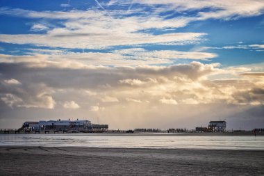 Sankt Peter-Ording, Almanya 'da dramatik gökyüzüne karşı gün batımında alçak gelgitte nemli evlerin panoramik manzarası ve ahşap kanalizasyon.