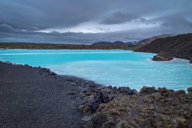 Mavi Göl 'ün parlak turkuaz jeotermal suyu İzlanda' da dramatik bulutlu bir gökyüzünün altında kara lav kayalarıyla çelişiyor.