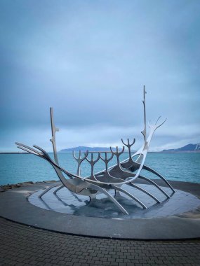 Reykjavik, İzlanda - 28 Mart 2022: Paslanmaz çelik Güneş Voyager heykeli, arka planda bulutlu gökyüzüne karşı okyanus ve dağlarla rıhtımda duruyor