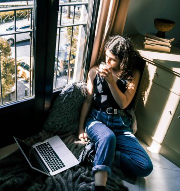 Pencerenin yanında dizüstü bilgisayarla çalışan genç bir kız.