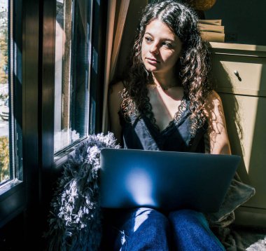 Pencerenin yanında dizüstü bilgisayarla çalışan genç bir kız.