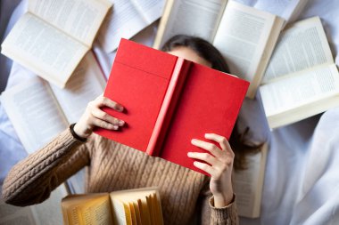 Yatakta kırmızı kitap okuyan bir kadın. Birçok açık kitap beyaz bir yatakta..