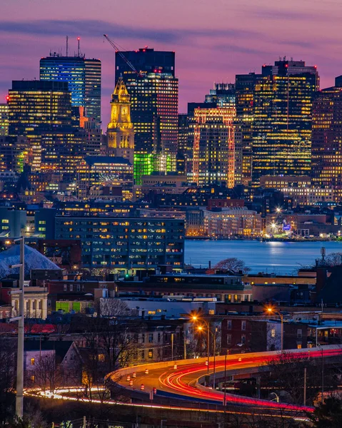 Boston city buildings and light trails under a pink sunset sky.