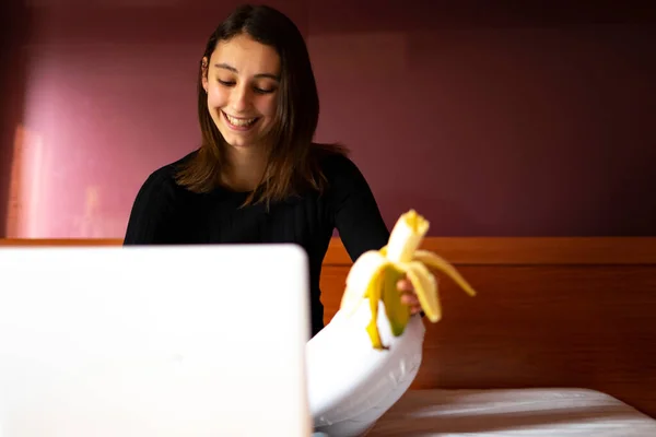 Bilgisayarla Yatakta Çalışan Muz Yiyen Bir Kadın Meyve Kavramı — Stok fotoğraf