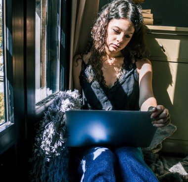 Pencerenin yanında dizüstü bilgisayarla çalışan genç bir kız.