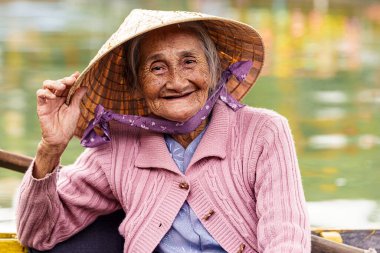 Mor kazaklı yaşlı Vietnamlı kadın gülümserken şapkasını tutuyor.