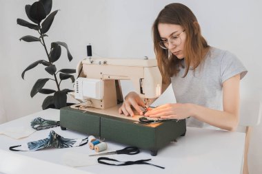 Gribi önlemek için evdeki dikiş makinesine koruyucu ilaç maskesi takan bir kız. Covid-19 beyaz arka plan ev işyeri olan odada.