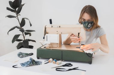 Gribi önlemek için evdeki dikiş makinesine koruyucu ilaç maskesi takan bir kız. Covid-19 beyaz arka plan ev işyeri olan odada.