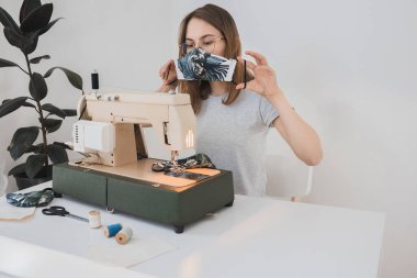 Gribi önlemek için evdeki dikiş makinesine koruyucu ilaç maskesi takan bir kız. Covid-19 beyaz arka plan ev işyeri olan odada.
