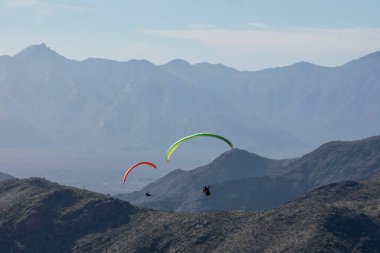 Paraşütçüler paraşütlerden sarkıyor dağların tepesinde rüzgarda süzülüyorlar.