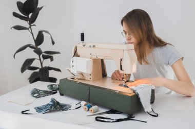 Gribi önlemek için evdeki dikiş makinesine koruyucu ilaç maskesi takan bir kız. Covid-19 beyaz arka plan ev işyeri olan odada.