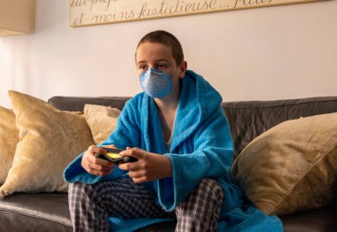 COVID-19 salgını sırasında yüz maskesi altında video oyunu oynayan genç bir çocuk.