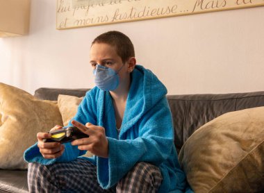 COVID-19 salgını sırasında yüz maskesi altında video oyunu oynayan genç bir çocuk.