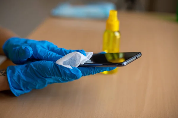 Woman's hand in blue gloves sanitizing cleaning smartphone mobile phone on wood table surface with wet wipes and alcohol