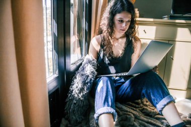 Pencerenin yanında dizüstü bilgisayarla çalışan genç bir kız.