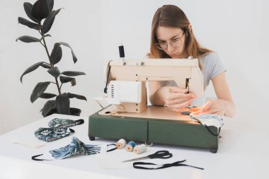 Gribi önlemek için evdeki dikiş makinesine koruyucu ilaç maskesi takan bir kız. Covid-19 beyaz arka plan ev işyeri olan odada.
