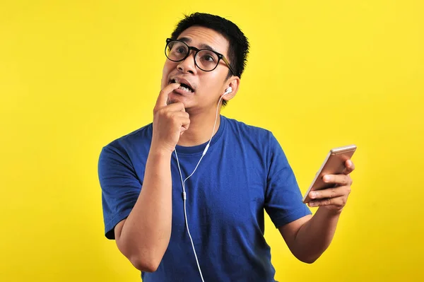 困惑的亚洲年轻人的画像 他用耳机在智能手机上咬着手指 听着现场音乐 背景是黄色的 — 图库照片