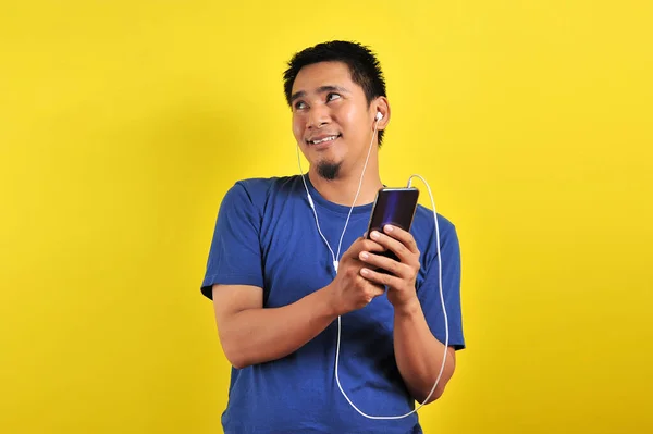 身穿休闲蓝色T恤 头戴耳机 听智能手机音乐的亚洲人 背景是黄色的 — 图库照片