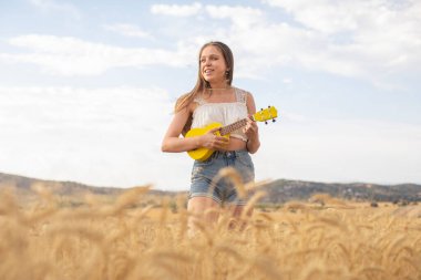 Gün batımında mısır gevreği tarlasında sarı ukulele çalan genç bir kadın.