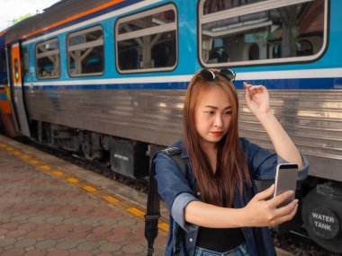 Asyalı kadınlar tren istasyonunda arka planda trenle seyahat ederken akıllı telefon kullanıyorlar. Genç Asyalı kadın tren istasyonunda akıllı telefona bakıyor..