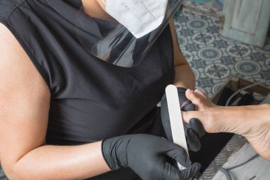 Covid19 sırasında maskeli ve koruyucu ekranlı müşteri için pedikür yapan bir güzellik uzmanı.