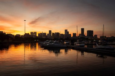 Beautiful sunset view to boats in local marina with yellow and orange reflections on the water and downtown buildings on the back, Rio de Janeiro, Brazil clipart
