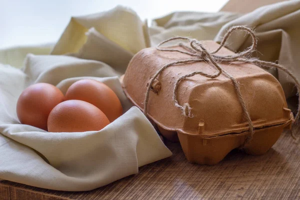 Delicate still life with three brown eggs on a light cloth as a nest and a egg carton packed with string on a kitchen board. Soft beige tones.Horizontal view.