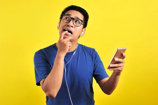 困惑的亚洲年轻人的画像 他用耳机在智能手机上咬着手指 听着现场音乐 背景是黄色的 — 图库照片