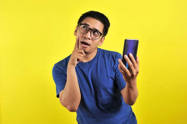Asiatischer Mann Mit Smartphone Macht Denkgeste Isoliert Auf Gelbem Hintergrund — Stockfoto