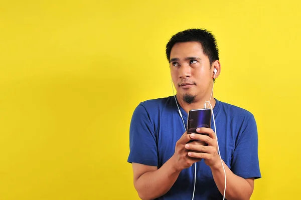 身穿休闲蓝色T恤 头戴耳机 听智能手机音乐的亚洲人 背景是黄色的 — 图库照片
