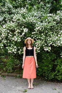 Yazlık şapkalı ve gözlüklü genç bir kadın çiçek açan ağaçların arasında yürüyor.